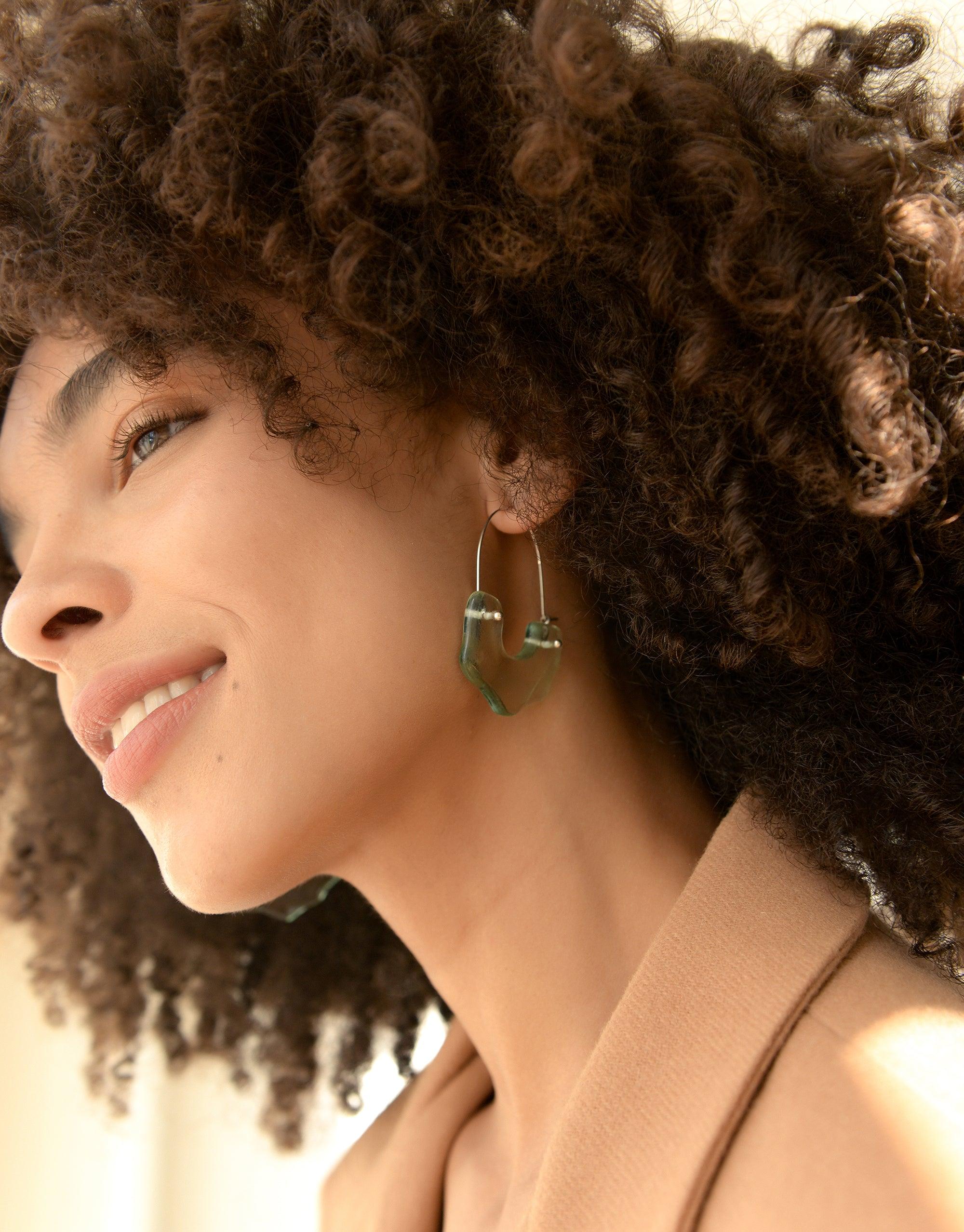 Close-up profile of a model with curly hair wearing unique arch-shaped hoop earrings, showcasing the Polar Arch Hoop Earrings from the Floating Glacier Collection. The earrings feature a distinctive eco-friendly design made from repurposed glass. CLED