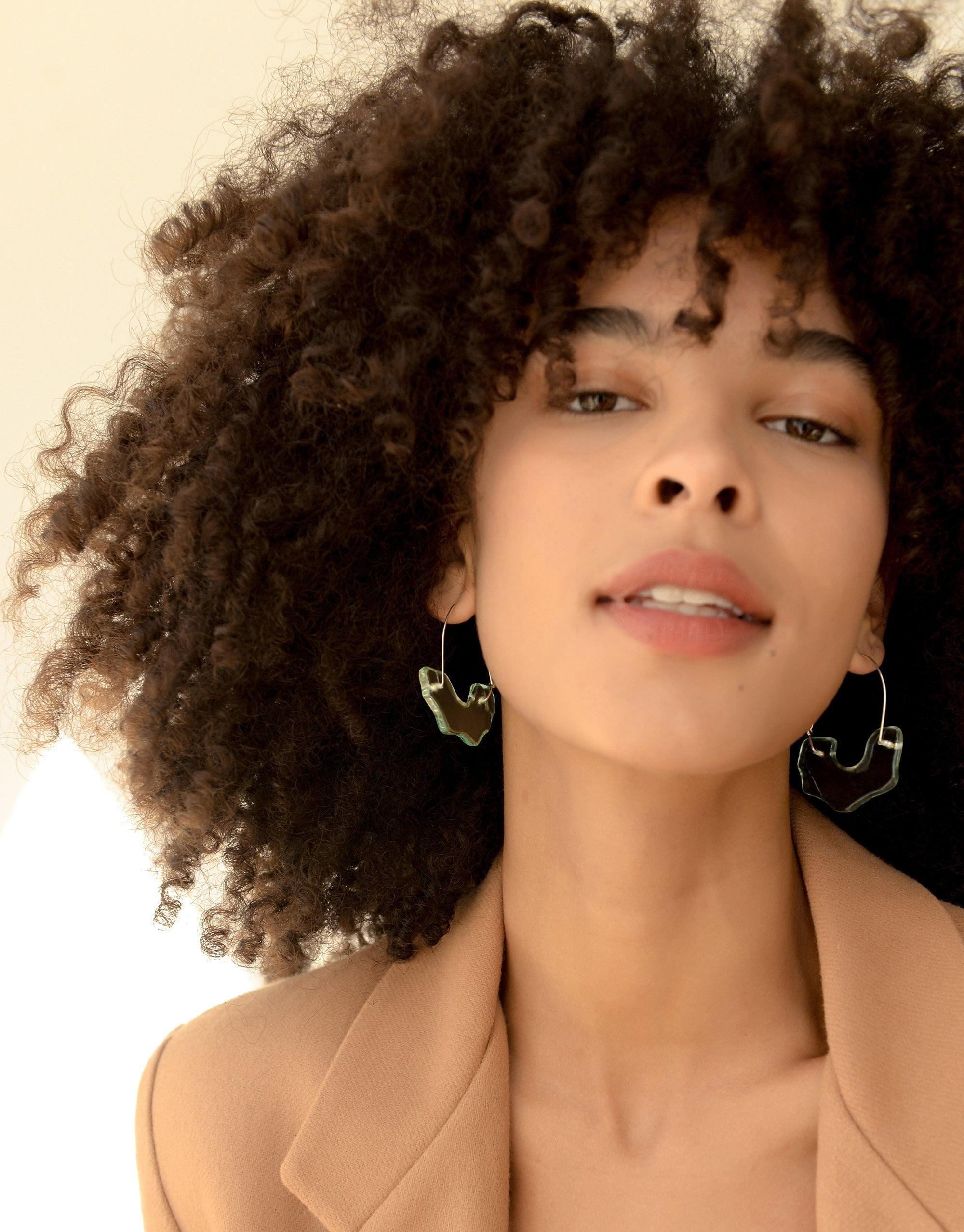 Close-up of model with curly hair wearing elegant arch-shaped hoop earrings, showcasing the unique design of Polar Arch Hoop Earrings. The earrings feature abstract lines inspired by icebergs, highlighting the eco-friendly and climate change awareness aspect of the Floating Glacier Collection by CLED.