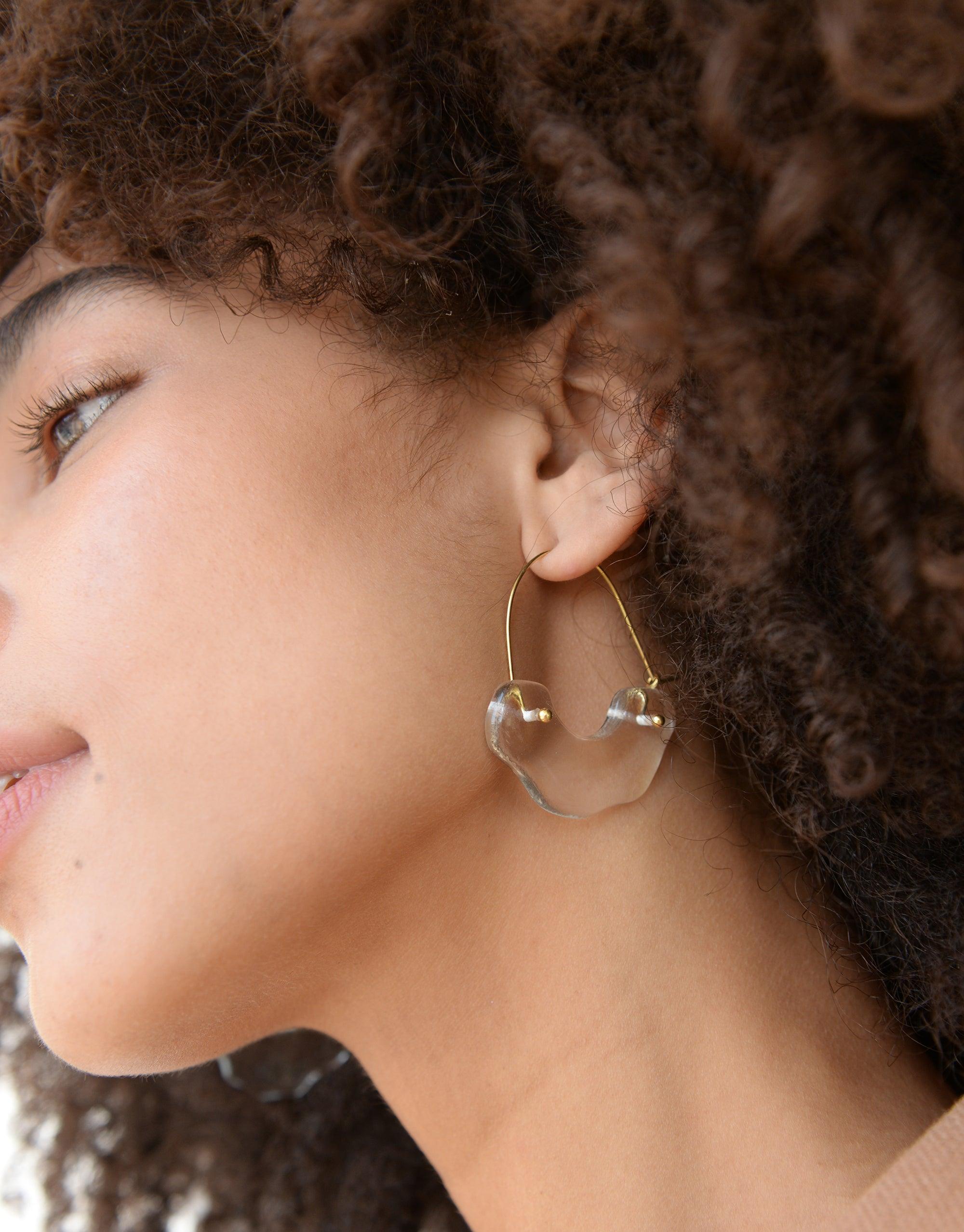 Close-up of elegant Polar Arch Hoop Earrings with curved glass design, worn by a model with curly hair, showcasing the earring's unique shape inspired by icebergs. Sustainable jewelry piece from the Floating Glacier Collection by CLED.
