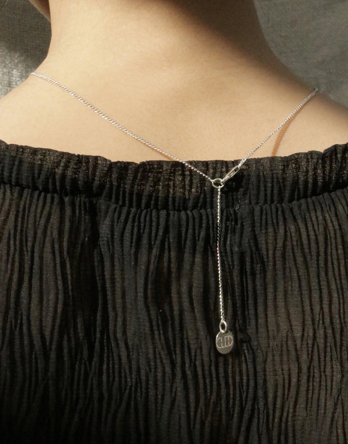 Close-up of a delicate silver chain necklace with a small loop pendant resting on a person's collarbone. The necklace contrasts beautifully against a black textured top. In The Loop Necklace by CLED showcases minimal elegance.