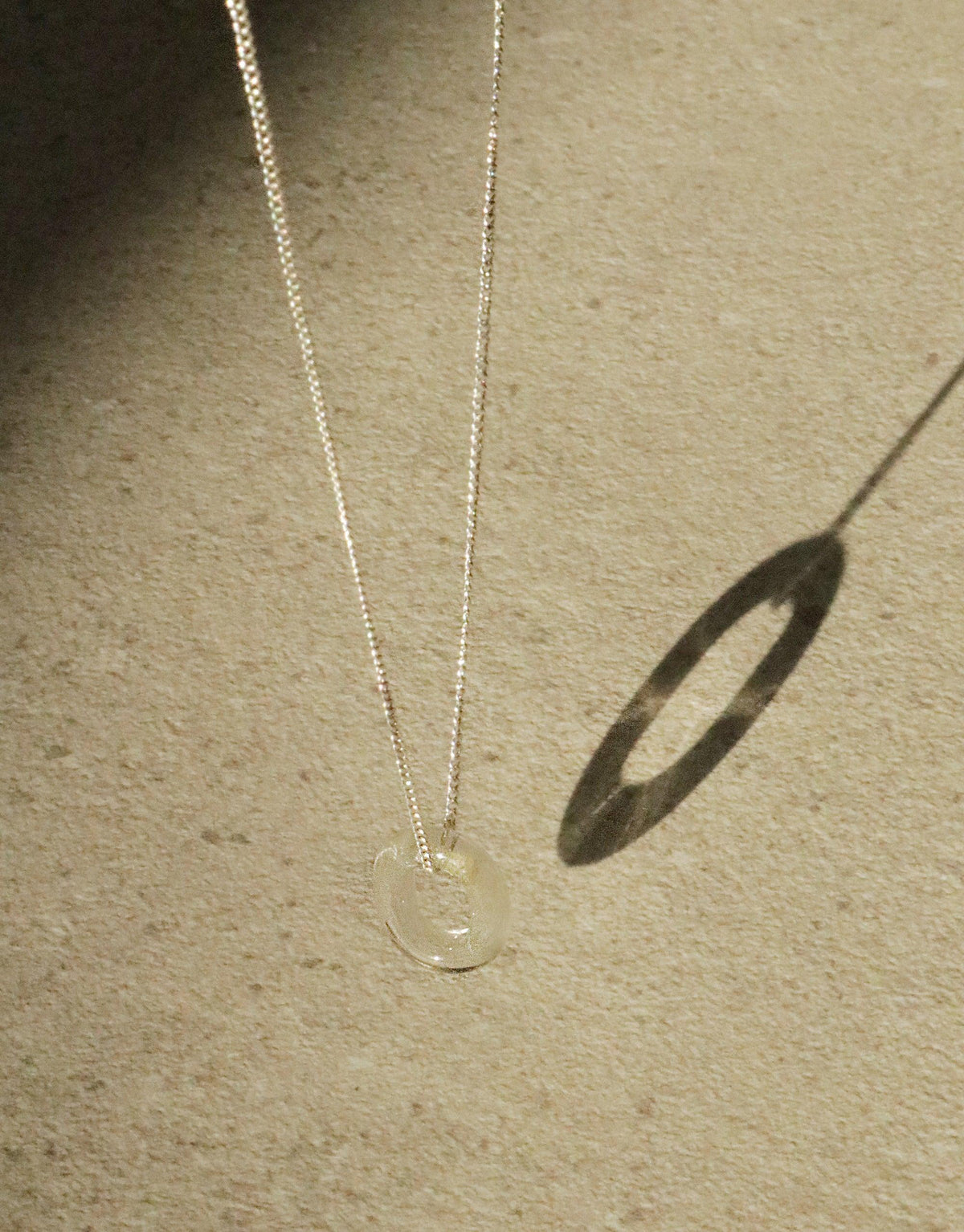 Delicate silver chain necklace with transparent eco-glass loop pendant, casting oval shadow on beige surface. Minimalist design showcases sustainable elegance in "In The Loop Necklace" by CLED.