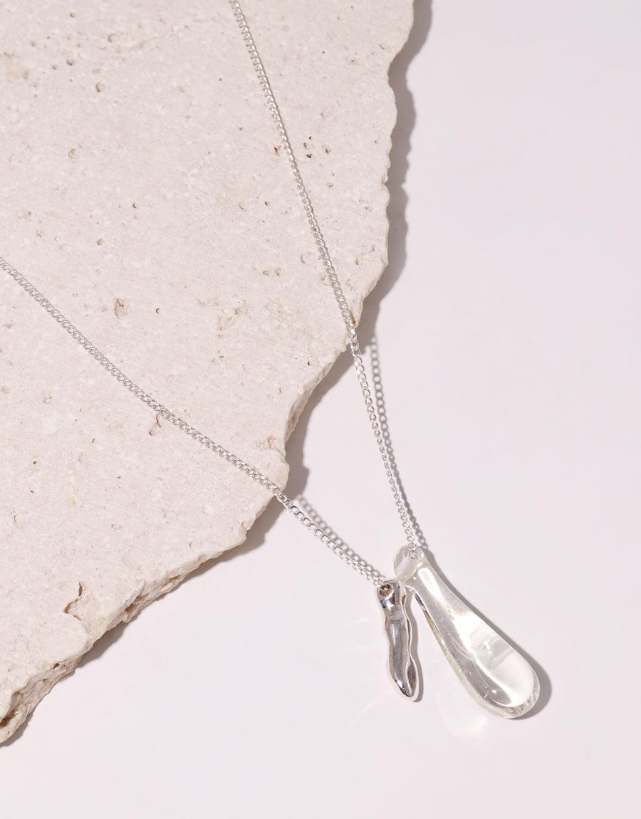 Delicate silver necklace with glass water drop pendant and wavy metal charm, showcasing eco-friendly design on textured background. Drop Counts Necklace by CLED emphasizes sustainability and elegance in jewelry craftsmanship.