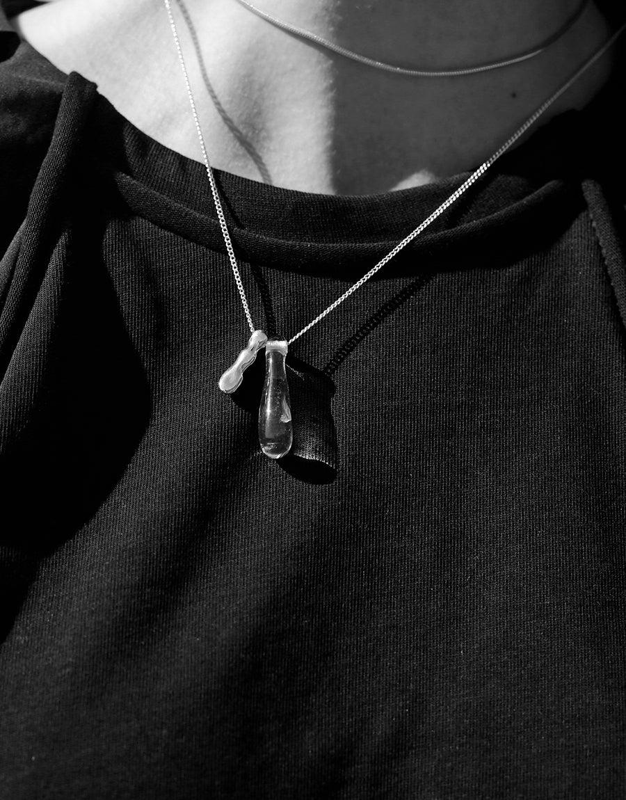 Close-up of Drop Counts Necklace featuring a delicate glass water drop pendant on a silver chain, worn against a black top. The pendant reflects light, emphasizing its eco-friendly design made from repurposed glass. Sustainable jewelry by CLED.
