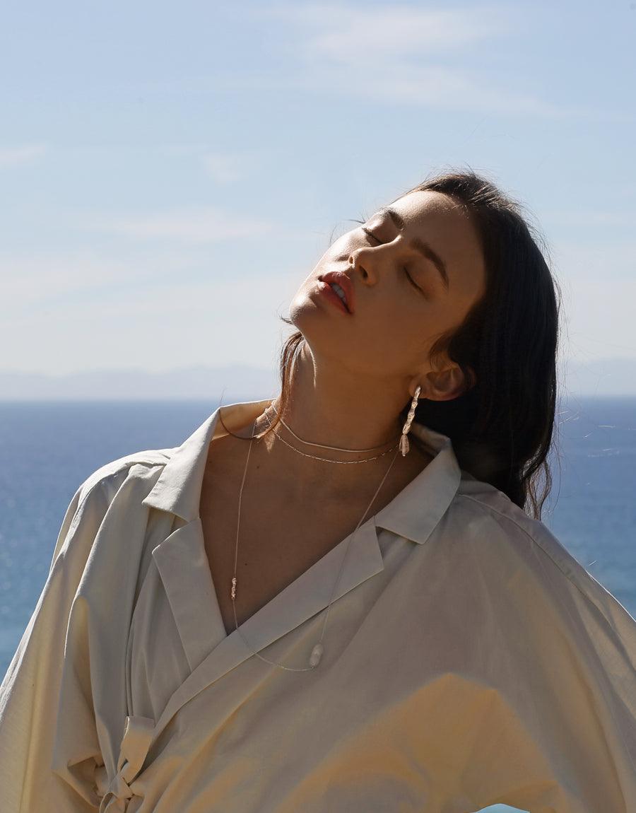Elegant model wearing Shoreline Long Necklace against ocean backdrop, showcasing delicate sterling silver chain with sustainable pearl and water drop pendant. Perfect for layering or individual wear. Handcrafted eco-friendly jewelry by CLED.