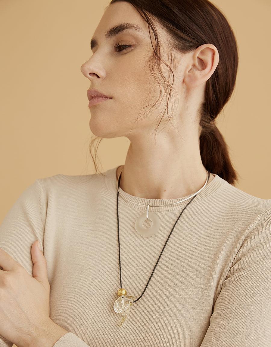 Profile view of a model wearing the Amorphous Long Cluster Necklace, showcasing its adjustable length and unique charms against a neutral beige sweater. The necklace features handcrafted glass gems and metallic accents, demonstrating its versatile and elegant design. CLED Amorphous Long Cluster Necklace.