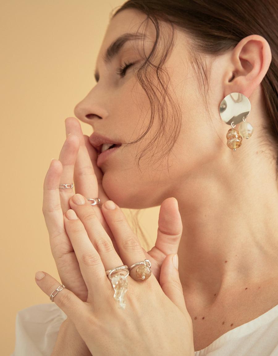 Profile view of a woman wearing multiple delicate rings, including the Collapsible Chain Ring | D, and statement earrings. Her hands are elegantly positioned near her face, showcasing the jewelry. Warm-toned background enhances the aesthetic. CLED