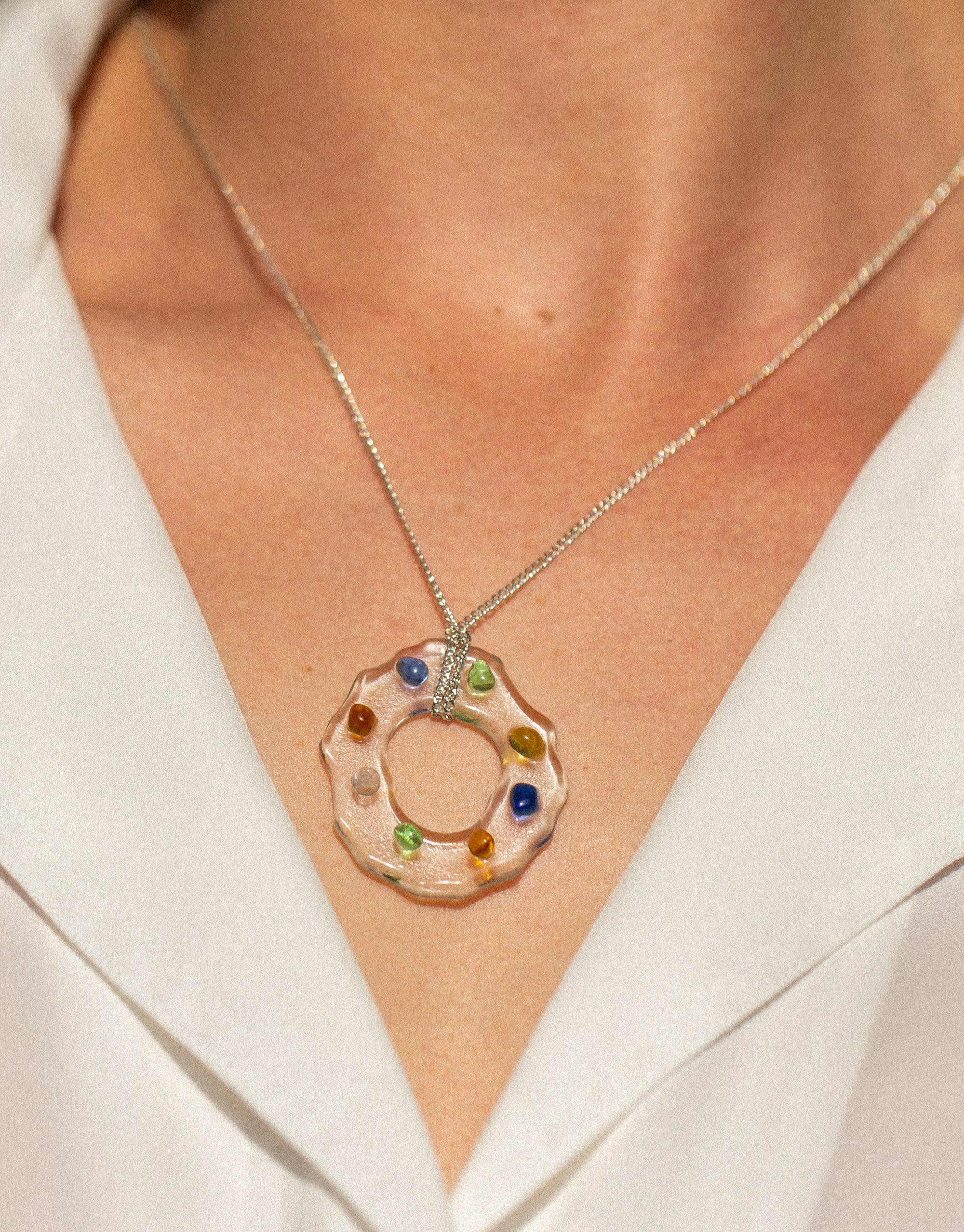 Close-up of High Tide Necklace in Spectrum featuring a circular pendant with colorful eco gems embedded in clear material, hanging on a delicate silver chain against a background of skin and white fabric. Sustainable jewelry by CLED.