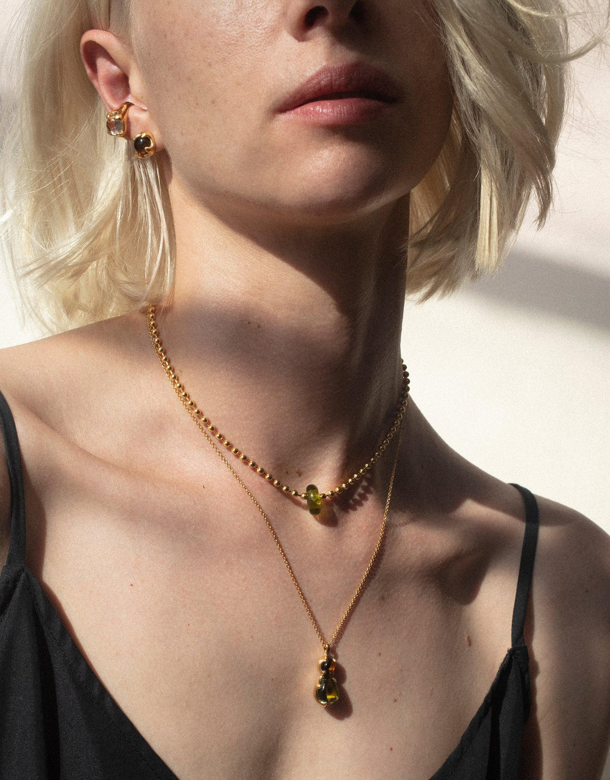 Close-up of a woman wearing layered gold necklaces featuring colorful eco gems, showcasing the Donut Ball Necklace. Sunlight highlights the prism-like effect of the jewelry, embodying the Rays of Light collection by CLED.