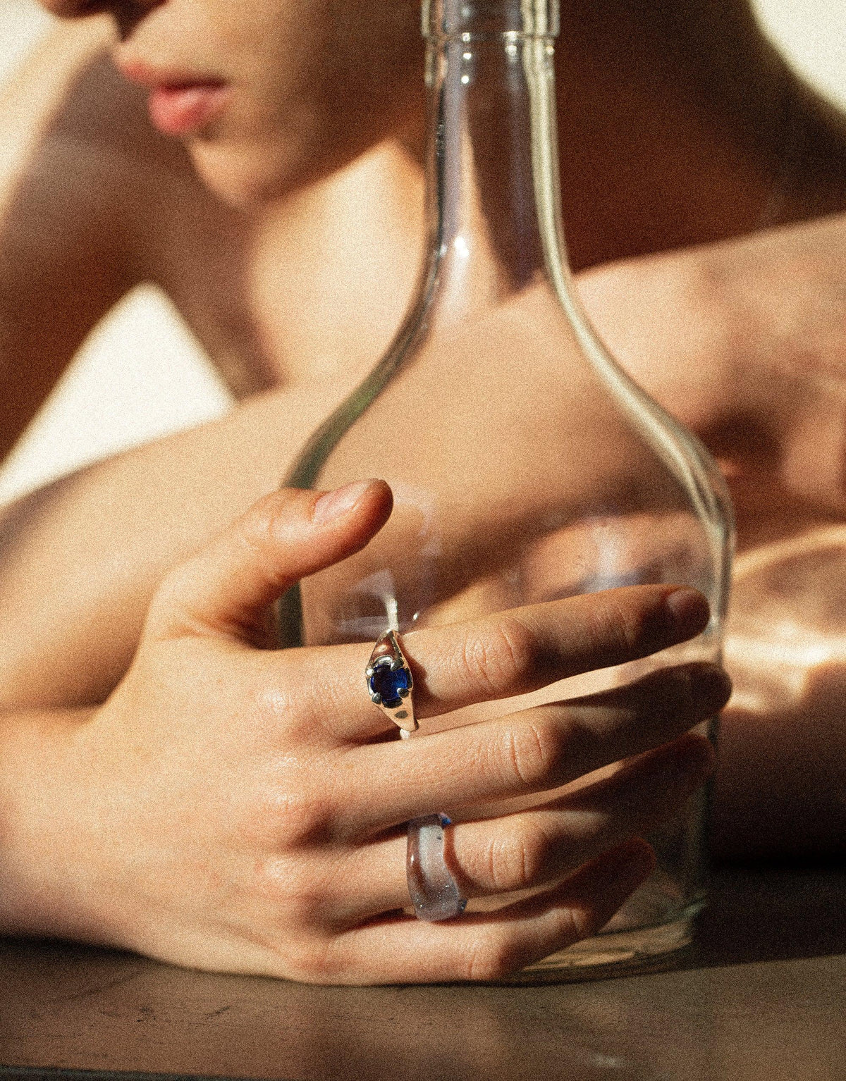 Close-up of hands holding a glass bottle, showcasing a blue gemstone ring on the finger. Soft, warm lighting highlights the jewelry and glass textures. Beam Ring by CLED.