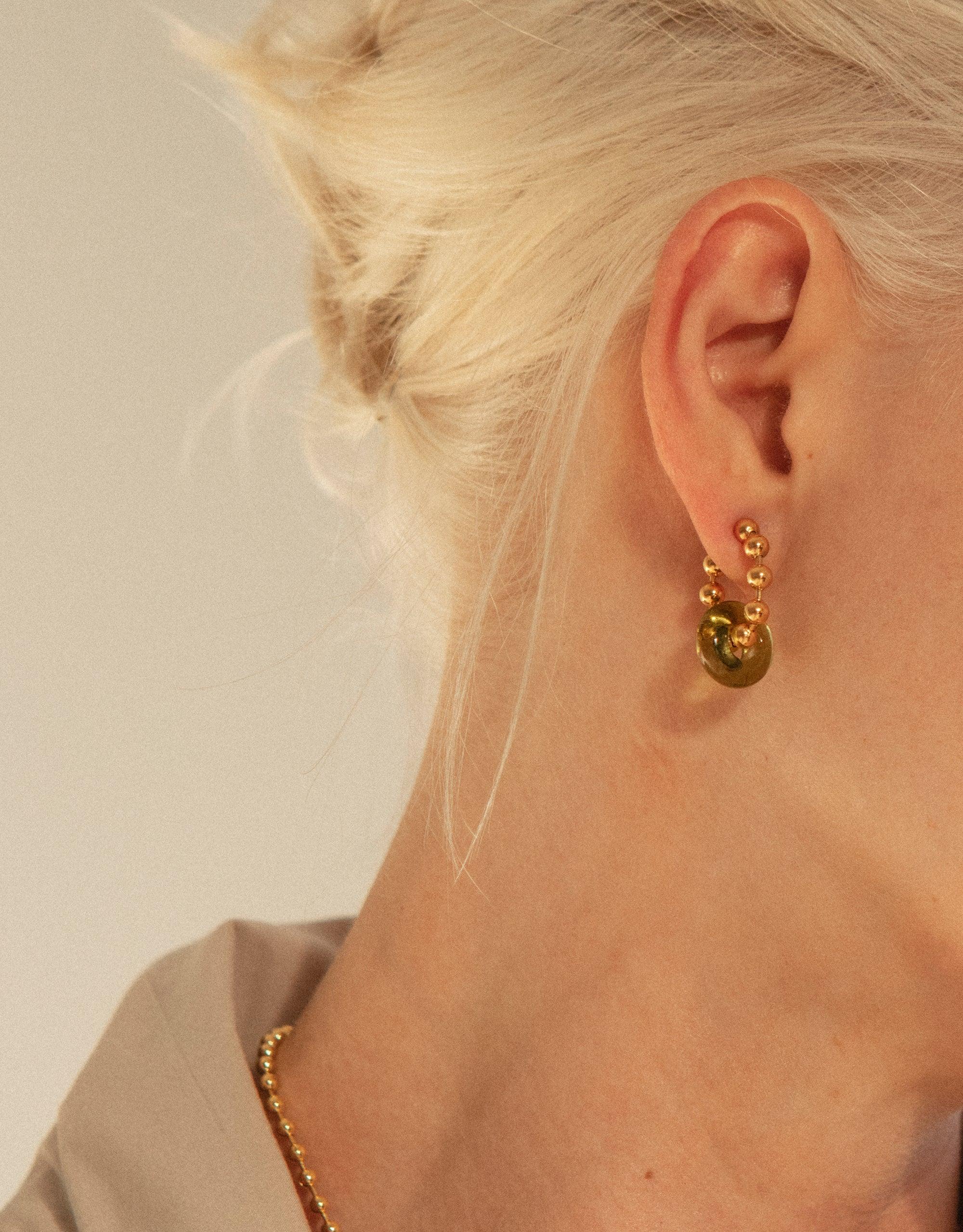 Close-up of a woman's ear wearing Donut Ball Hoop Earrings, featuring a gold-plated hoop with a green eco gem ball. The earring reflects light, showcasing the prismatic colors of the CLED upcycled glass jewelry. Donut Ball Hoop Earrings by CLED.