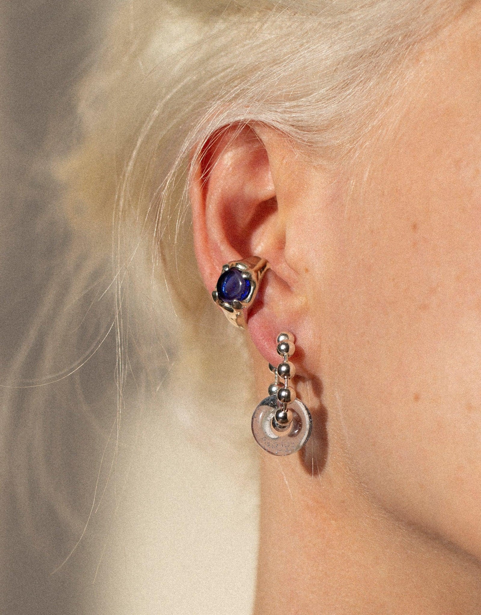Close-up of an ear wearing a shimmering blue gemstone stud and a delicate dangling silver earring, showcasing the unique Beam Ear Cuff design. Elegant jewelry against pale skin and blonde hair, highlighting the intricate craftsmanship of CLED's eco-friendly, handmade accessories.