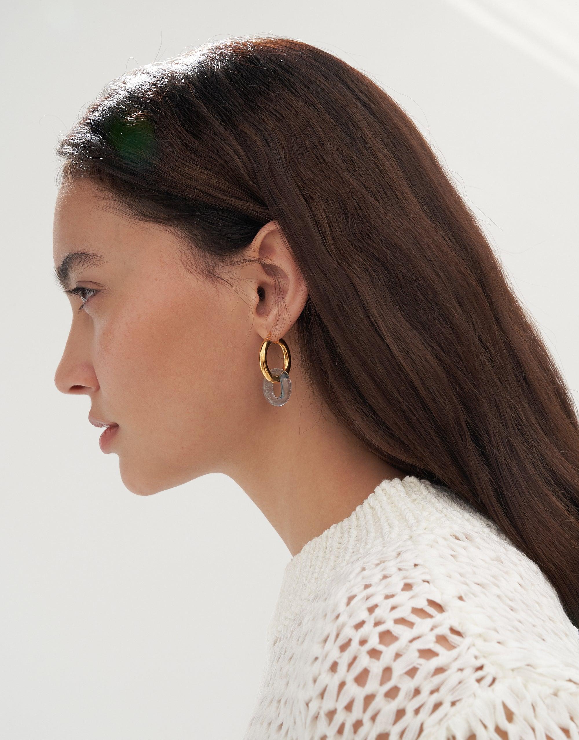 Side profile of a woman with long dark hair wearing a white knit sweater, showcasing large gold hoop earrings with a small eco gem pendant. The earrings exemplify the elegant design of In The Loop Hoops | Large by CLED.